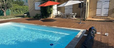 A swimming pool with a wooden deck, outdoor furniture, and a building with a tiled roof.