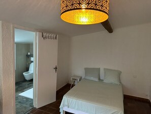 A bedroom with a bed, a nightstand, a lamp, and a bathroom visible through an open door.