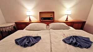 Un lit double avec une parure de lit à rayures, deux oreillers, deux serviettes pliées, une table de chevet en bois avec une radio et deux lampes fixées au mur.