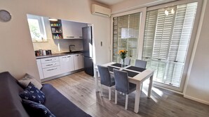 Un salon moderne comprenant un canapé, une table à manger avec des chaises, une cuisine avec des meubles blancs et une porte-fenêtre coulissante donnant sur l’extérieur.
