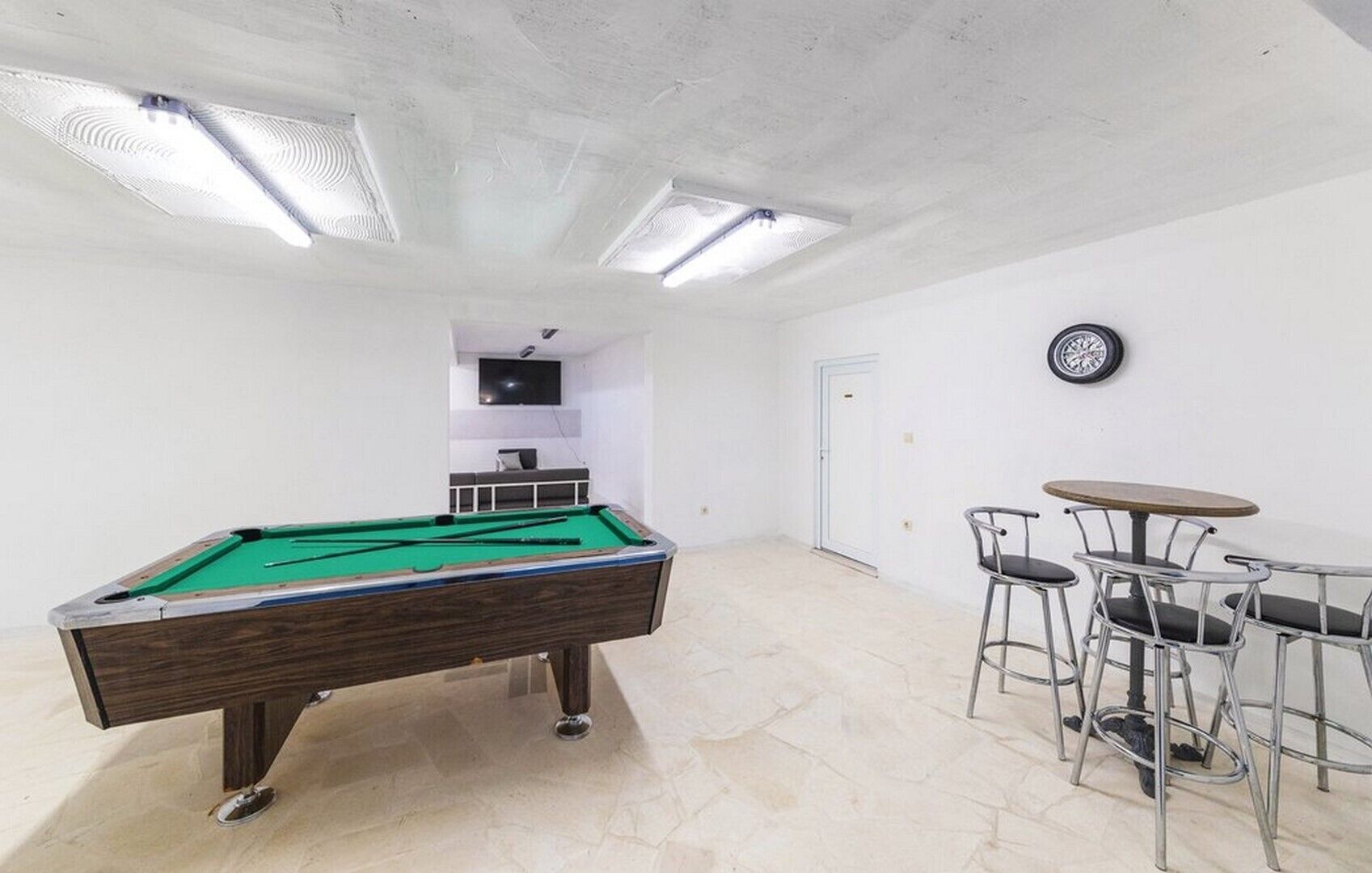 Een pooltafel met een groene viltbekleding, een klein zithoekje met metalen stoelen en een aan de muur bevestigde televisie.