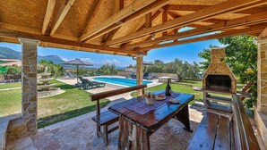 Un espacio al aire libre cubierto con un banco de piedra, una mesa con comida, un horno de piedra y una piscina al fondo.