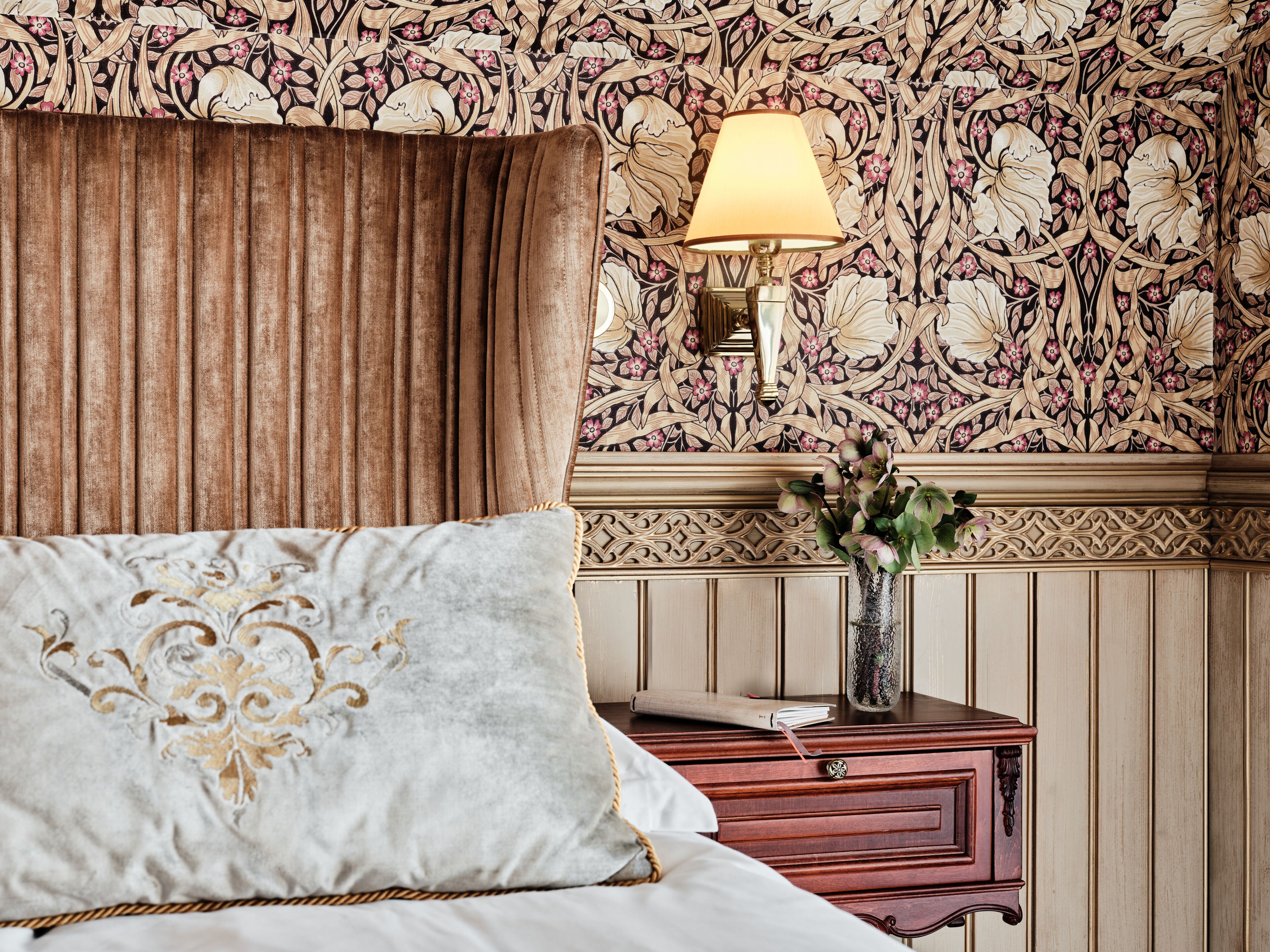 A bedroom with a bed, a nightstand, a lamp, and a floral wallpaper.