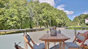 Una terraza con mesa y sillas, rodeada por una barandilla, con vista a una masa de agua y abundante vegetación.