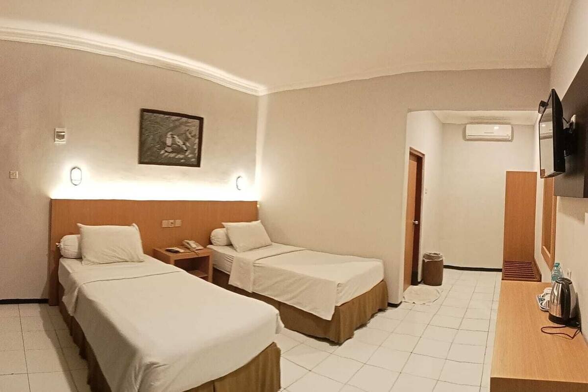 A hotel room with two single beds, a small desk with a telephone, a mirror, and a wall-mounted air conditioning unit.