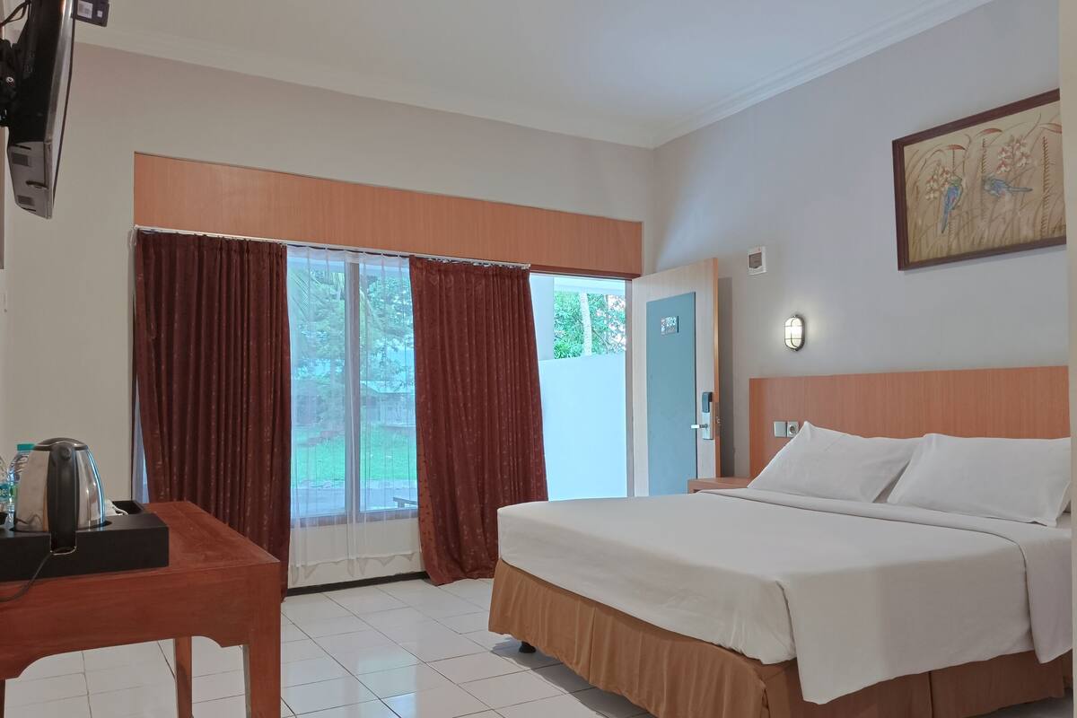 A hotel room with a bed, a desk with a kettle, curtains, a door, and a painting on the wall.