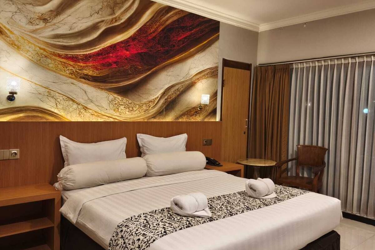 A hotel room with a large bed, a wooden nightstand, a small table, a chair, and a wall with a decorative mural.