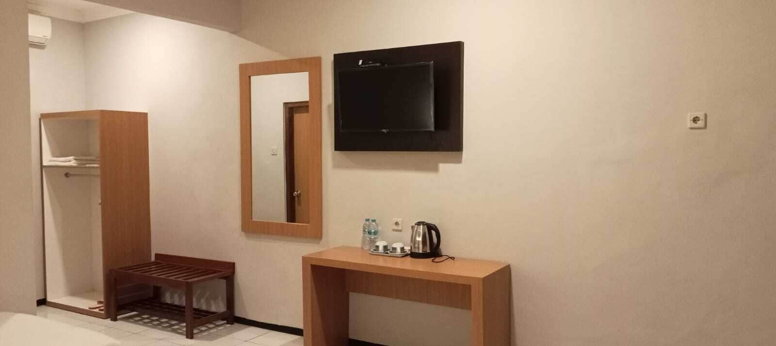 Habitación de hotel con un banco de madera, un espejo, un televisor de pantalla plana y una pequeña mesa con una tetera y botellas.