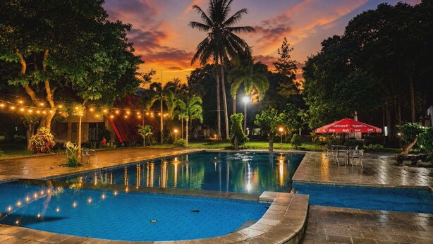 Una piscina con luces colgantes, palmeras y una puesta de sol al fondo.