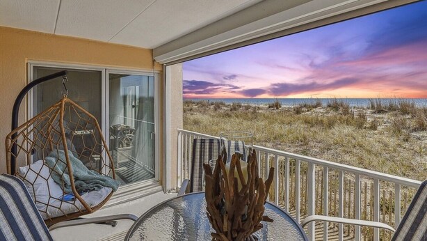 Un balcón con puerta de vidrio, una silla colgante, una mesa con un florero y vista a la playa al atardecer.