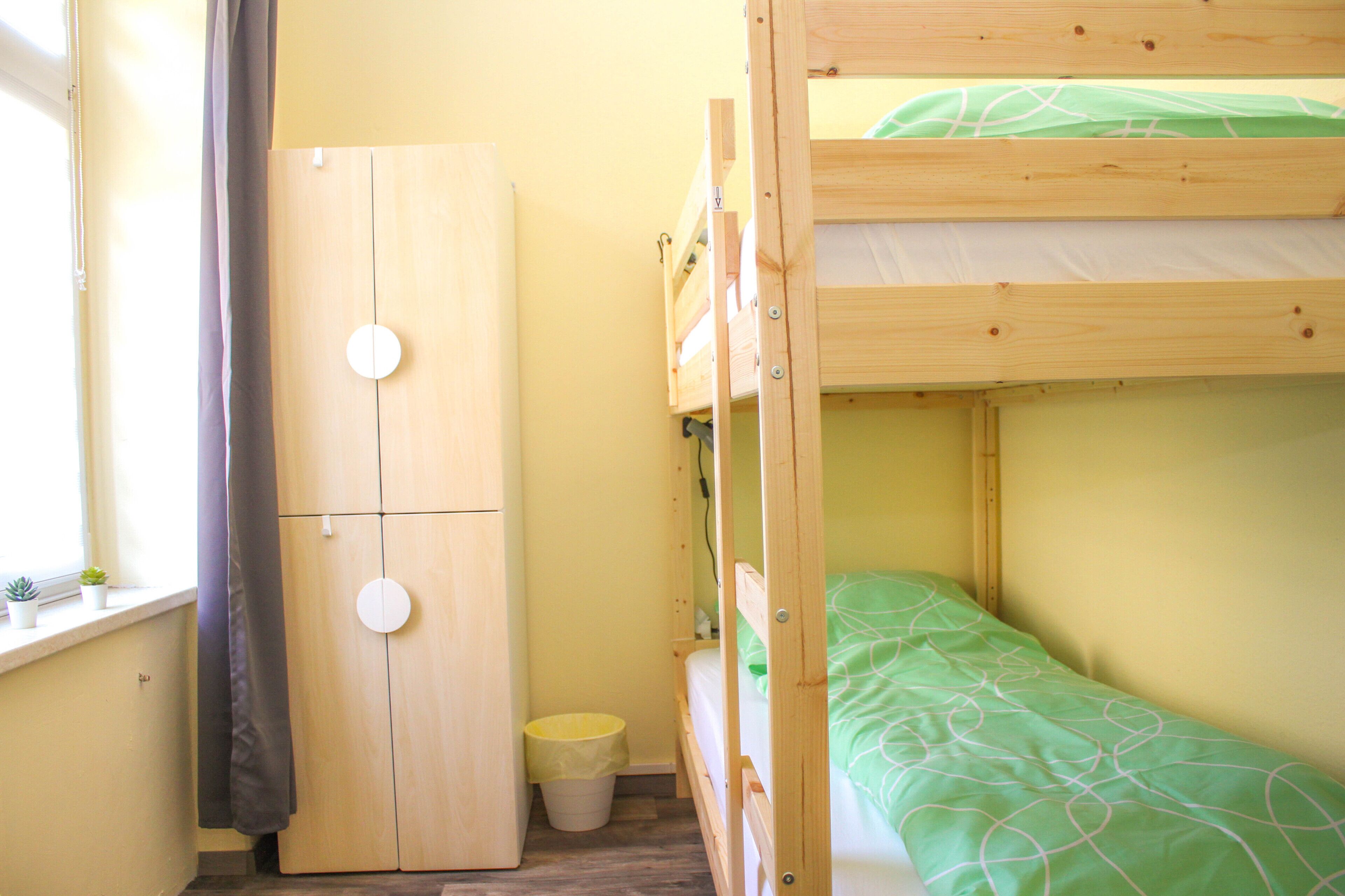 A bunk bed with a green patterned bedspread, a wooden wardrobe, and a small trash bin.
