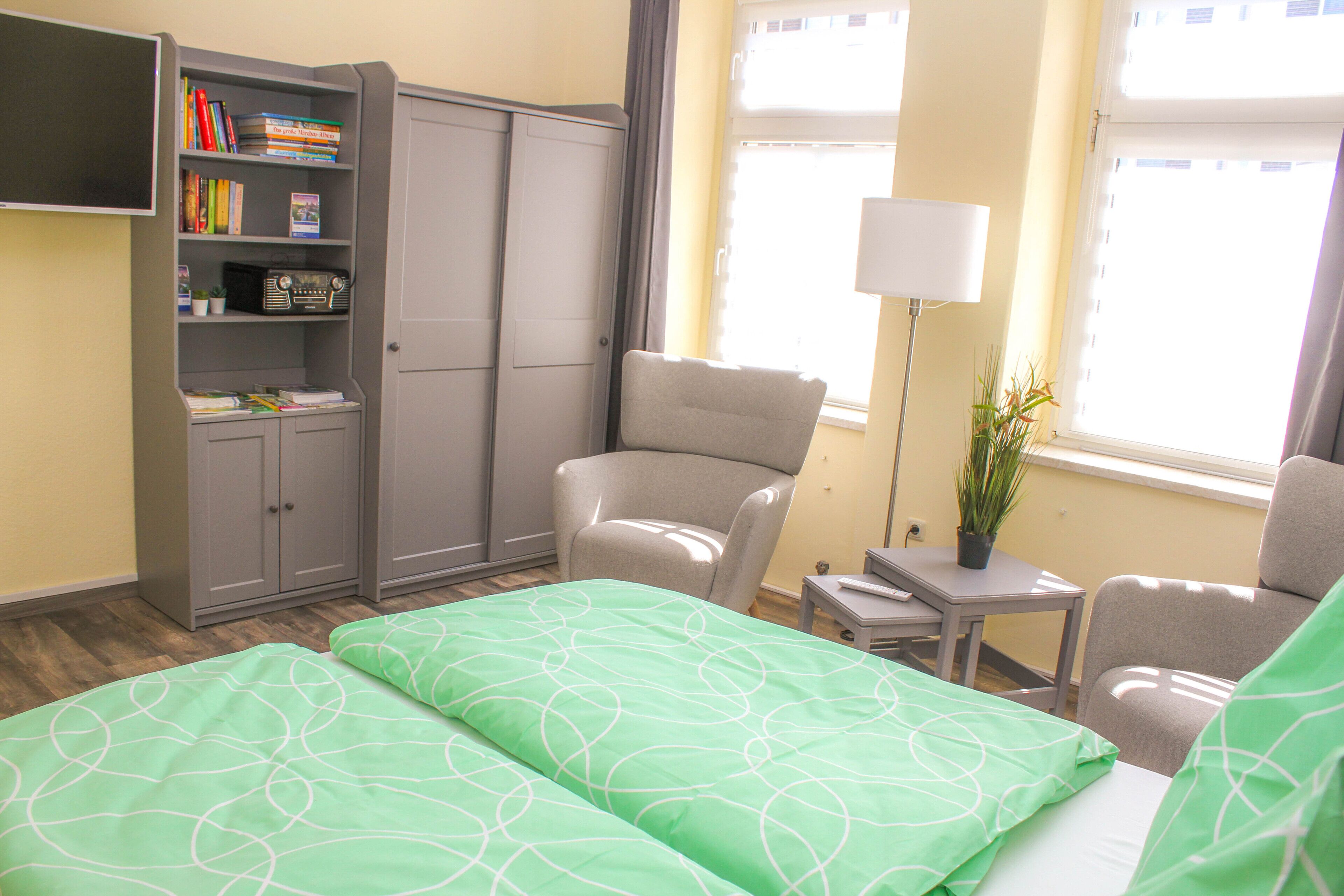 A hotel room with a bed, two chairs, a bookshelf, a desk, and a lamp.