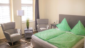 Una habitación de hotel con una cama, dos sillones, una mesita con lámpara y una planta en maceta.