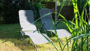 Dos sillones de exterior con estructura metálica y tapizado gris, ubicados sobre un césped con una valla de madera y abundante follaje al fondo.