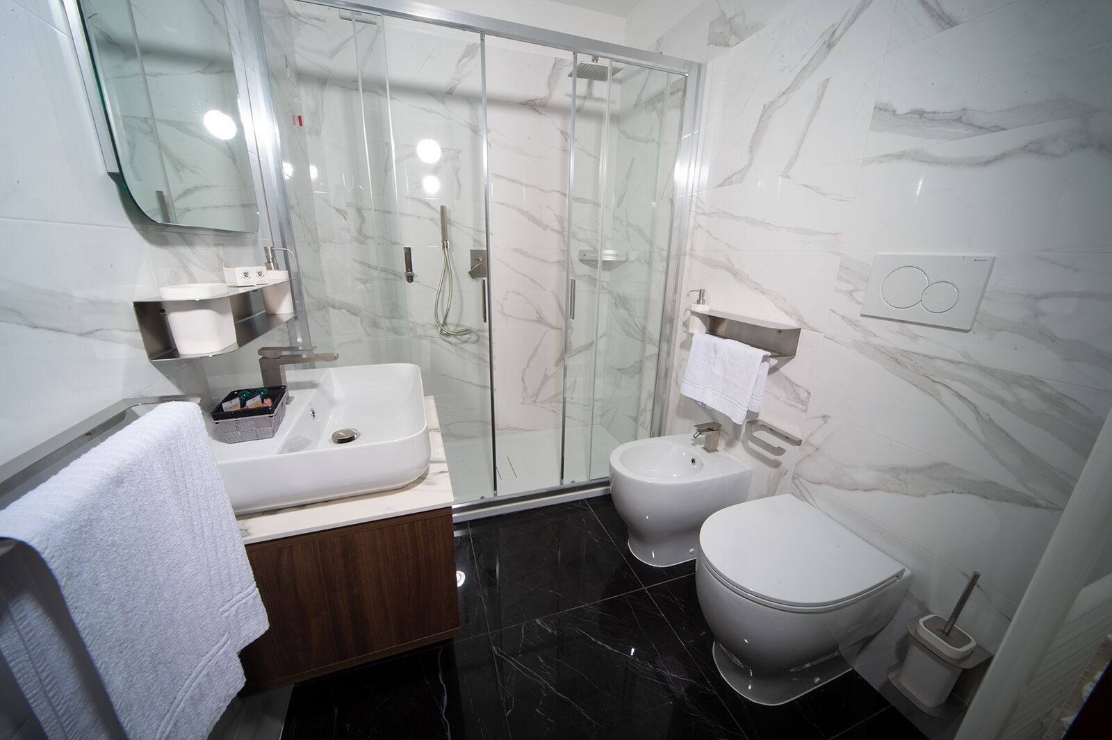 Un baño moderno con ducha envuelta en vidrio, un lavamanos blanco, un inodoro y suelo de baldosas de mármol.