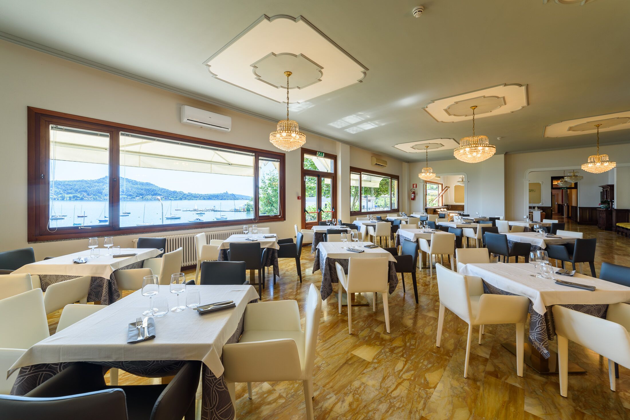 Un restaurante con varias mesas y sillas, amplias ventanas con vista al mar y luminarias decorativas en el techo.