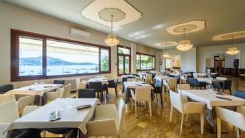 Un restaurante con varias mesas y sillas, amplias ventanas con vista al mar y luminarias decorativas en el techo.