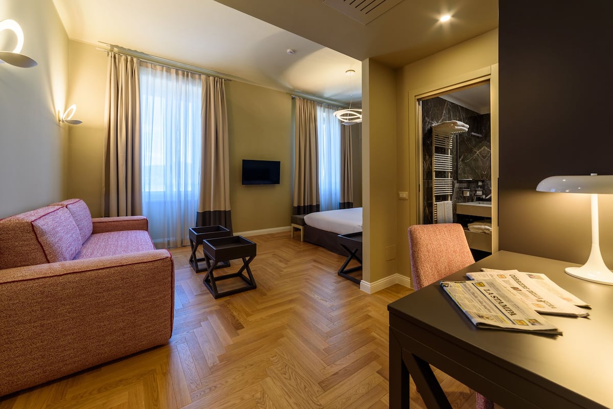 A hotel room with a sofa, a bed, a desk with newspapers, a TV, and a bathroom visible through an open door.