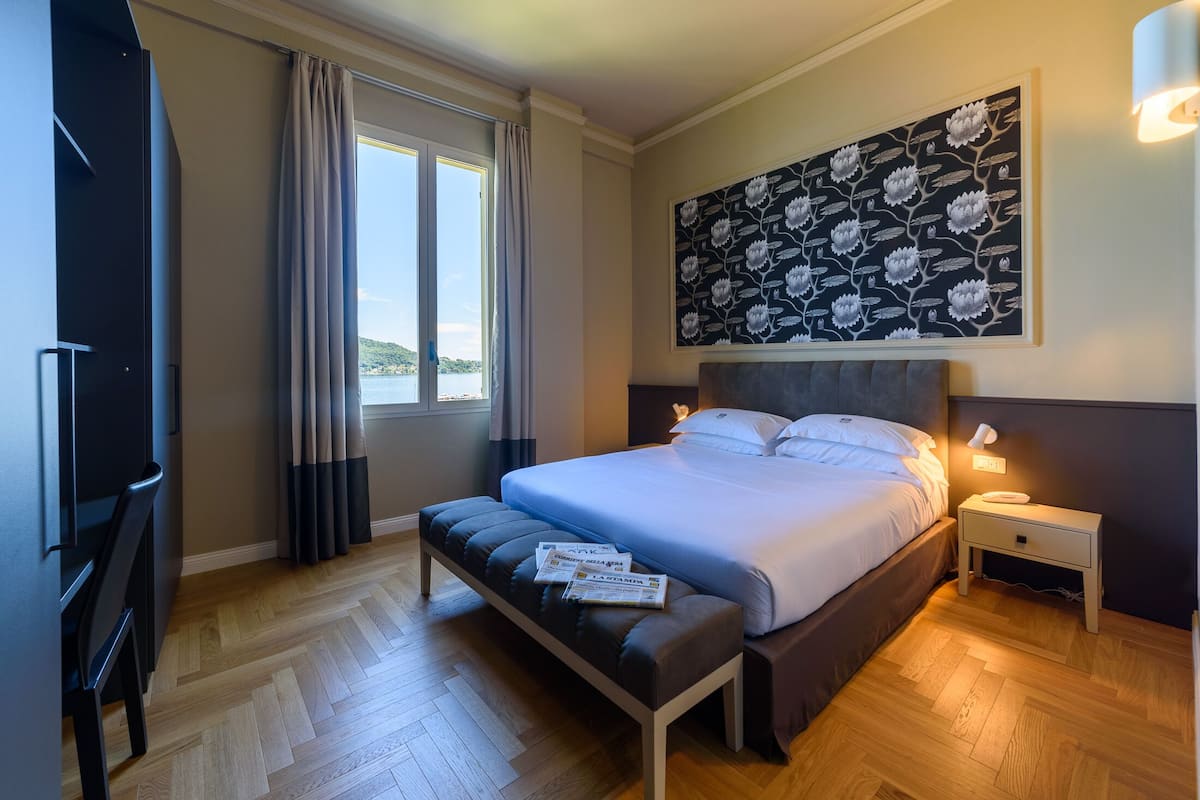 A bedroom with a large bed, a bench with magazines, a nightstand with a lamp, and a view of the sea through the window.
