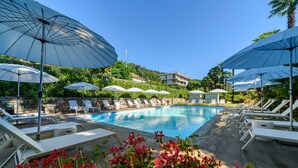 Zona de piscina con sillones de descanso y sombrillas, rodeada de vegetación y una colina.