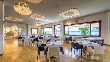 Un'ampia sala da pranzo con diversi tavoli e sedie, ampie finestre che offrono vista sull'esterno e lampadari decorativi.