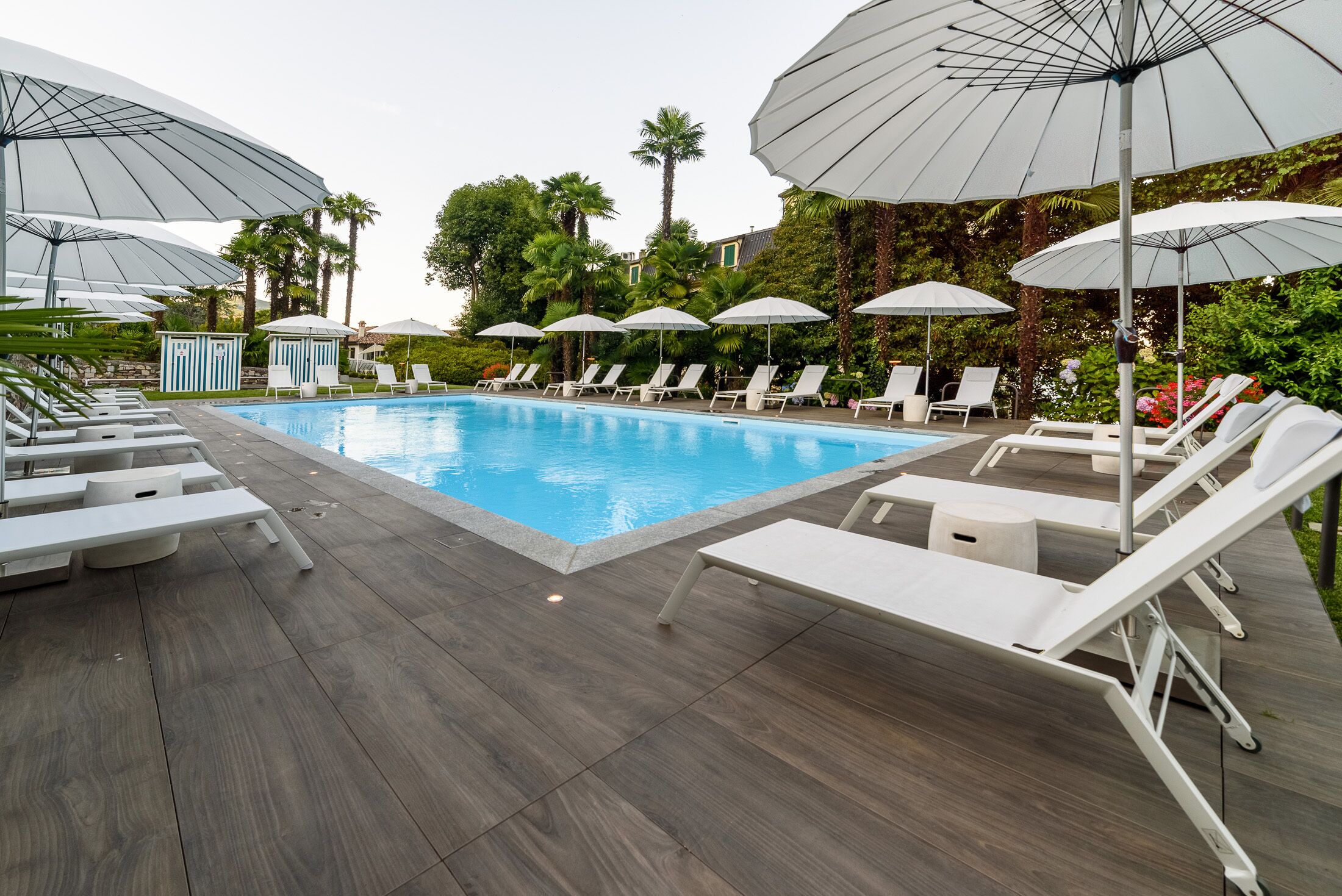 Piscina con sillas de descanso y sombrillas, rodeada de vegetación y una terraza de madera.