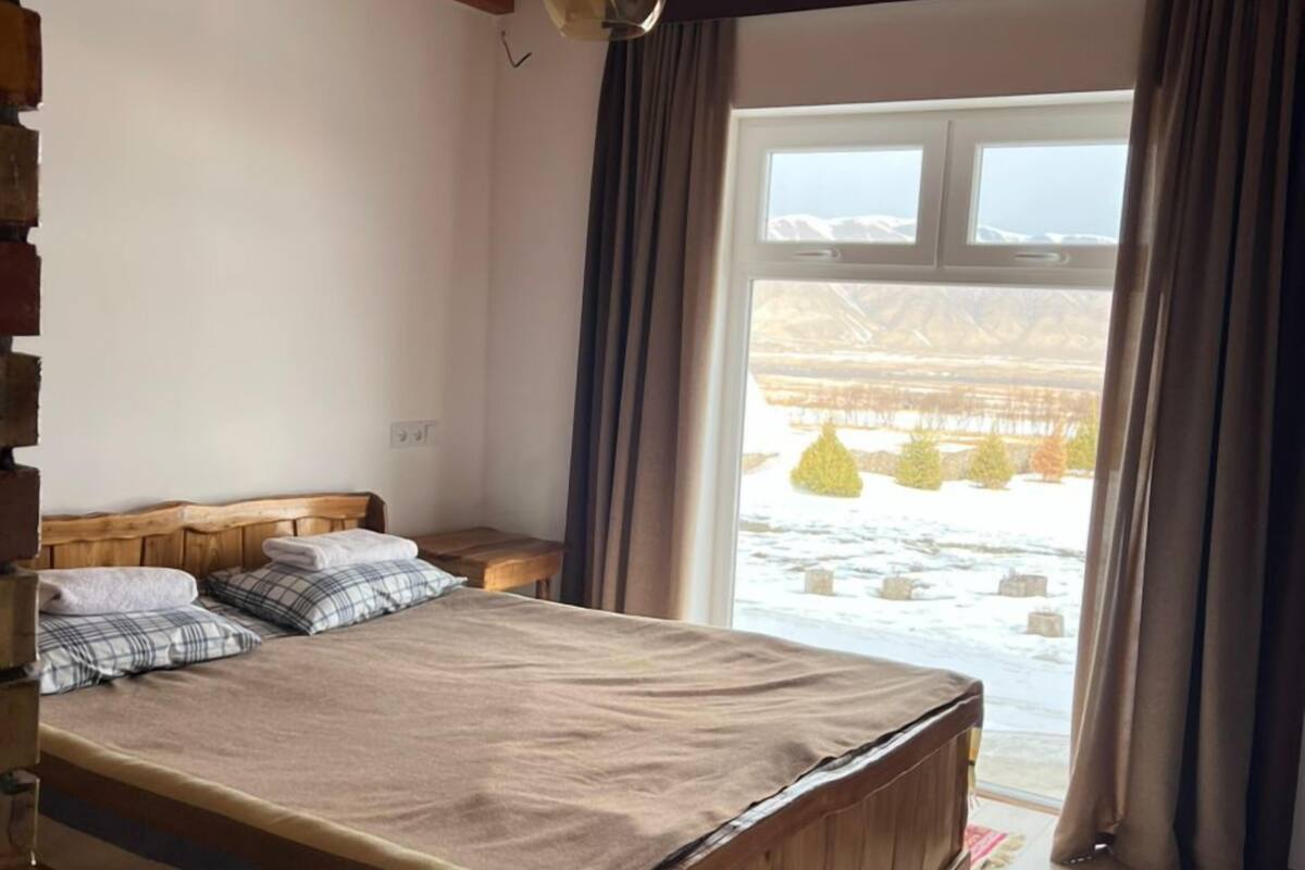 A wooden-framed bedroom with a large bed, a window with curtains, and a view of a snowy landscape.