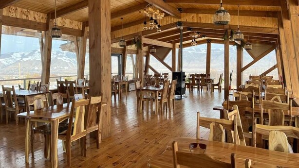Een ruime houten eetruimte met hoge plafonds en grote ramen met uitzicht op de bergen. Tafels en stoelen staan klaar voor een maaltijd.