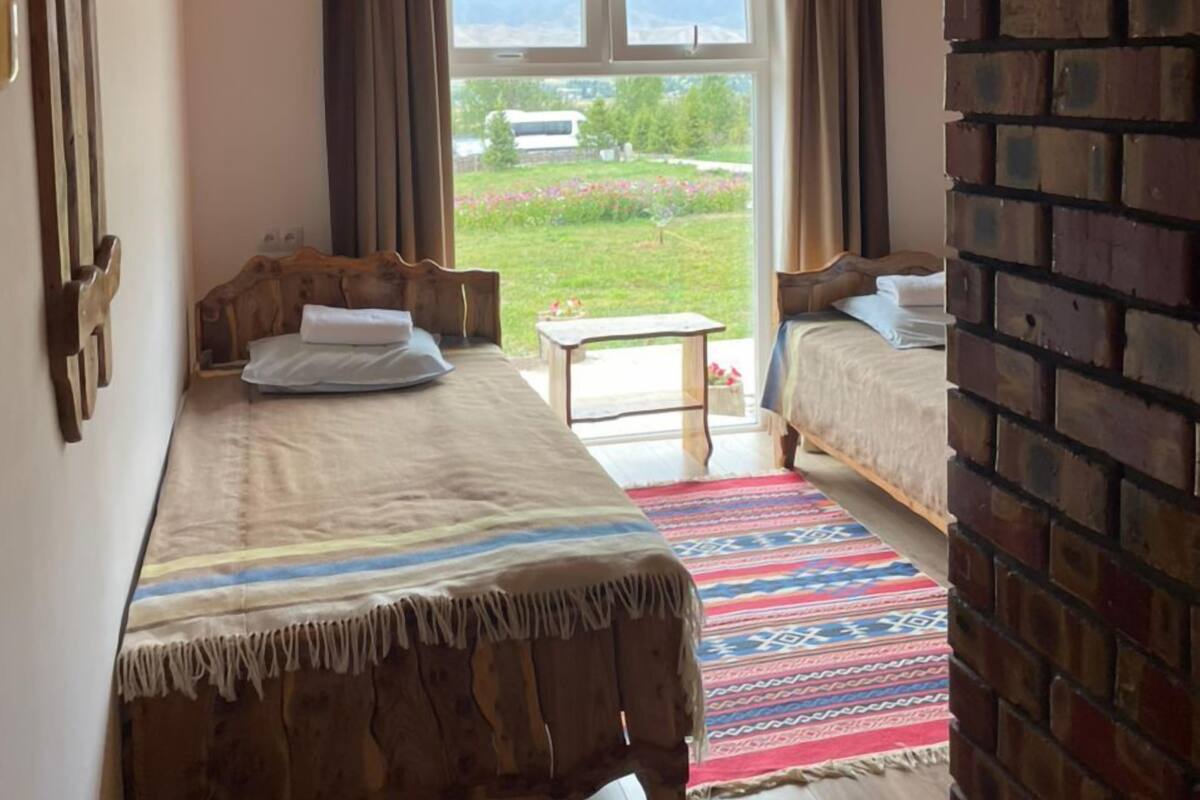 A bedroom with two beds, a wooden ceiling, a brick wall, and a view of a green landscape through a window.