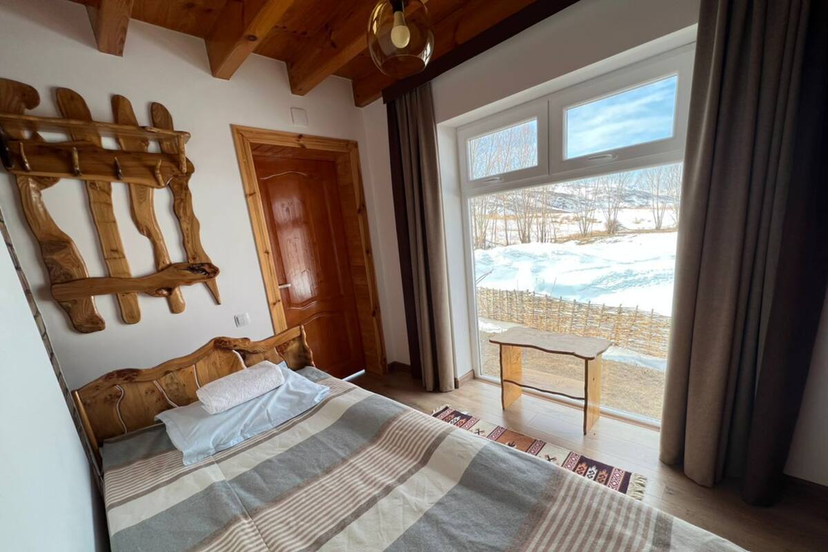 A bedroom with a wooden headboard, a large bed with a striped blanket, a wooden bench by a window, and a view of a snowy landscape.