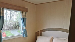 Un lit bien fait, avec du linge de lit blanc et un plaid gris, une tête de lit en bois et une fenêtre avec des rideaux.