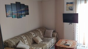 Sala de estar con un sofá, un televisor de pantalla plana montado en la pared, una mesita con un control remoto y un cuadro de un atardecer.