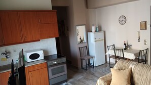 Cocina compacta con microondas, horno y armarios de madera. Hay un comedor con mesa y sillas, y un sofá en la sala. Se ve también un reloj y una puerta.