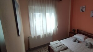Un dormitorio con cama, ventana con cortinas traslúcidas, mesita de noche y dos cuadros colgados en la pared.