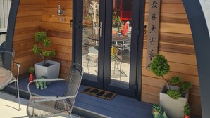 Una estructura de madera con techo curvo, una puerta y un pequeño patio con sillas y plantas en macetas.