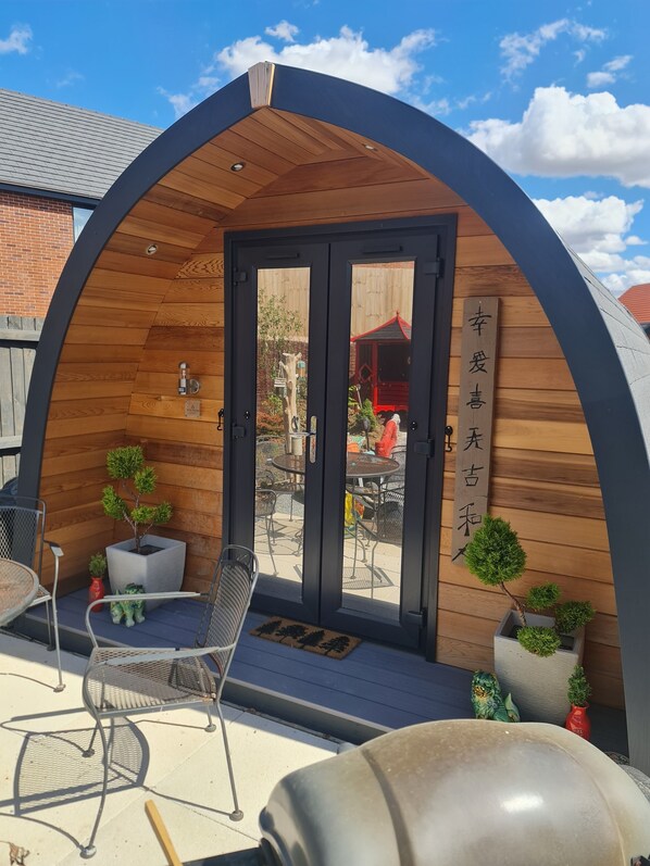 Una estructura de madera con techo curvo, una puerta y un pequeño patio con sillas y plantas en macetas.
