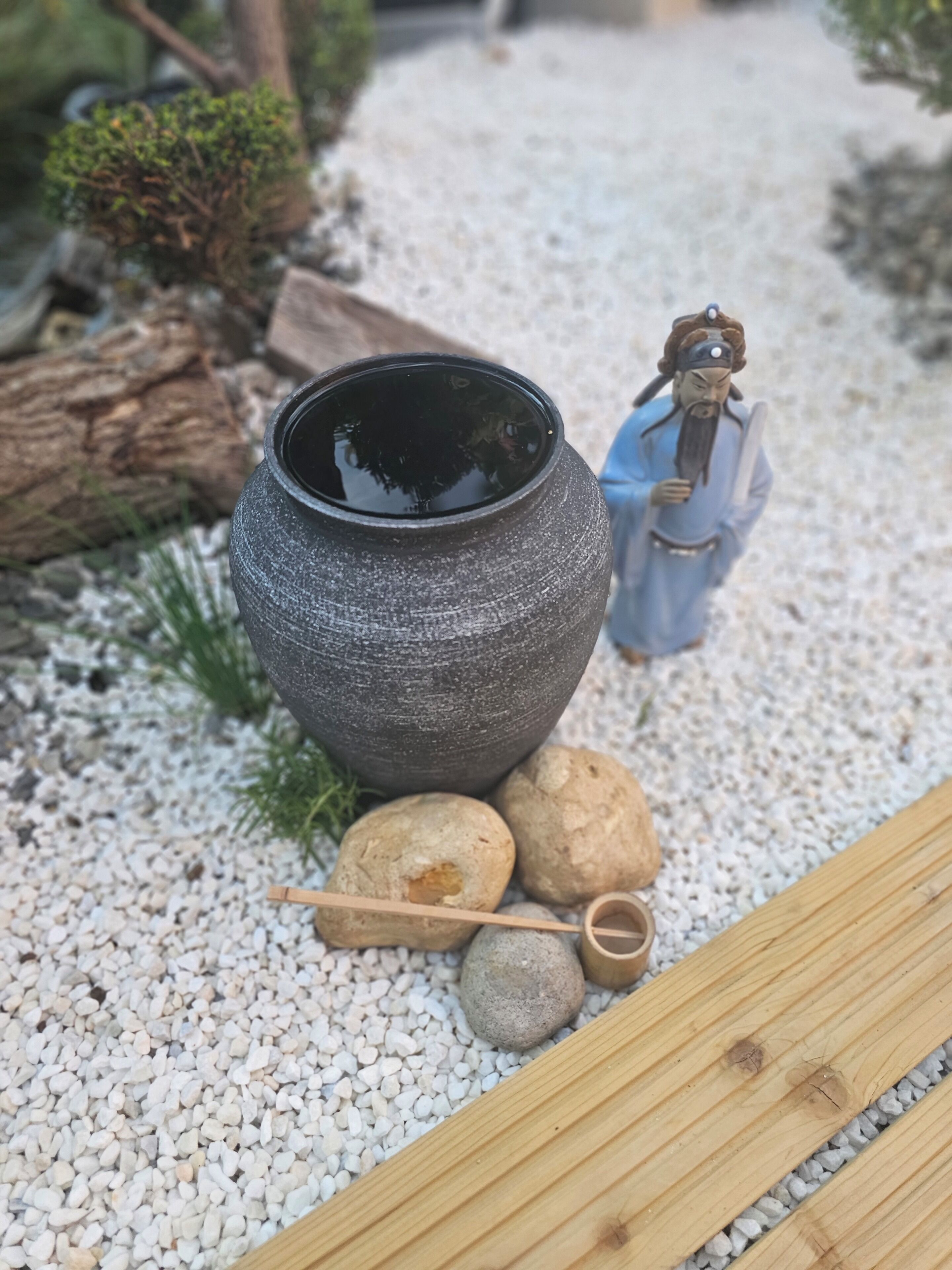 Een rustieke buitenomgeving met een grote terracotta pot, een miniatuurbeeldje en een houten dienblad op een grindpad.