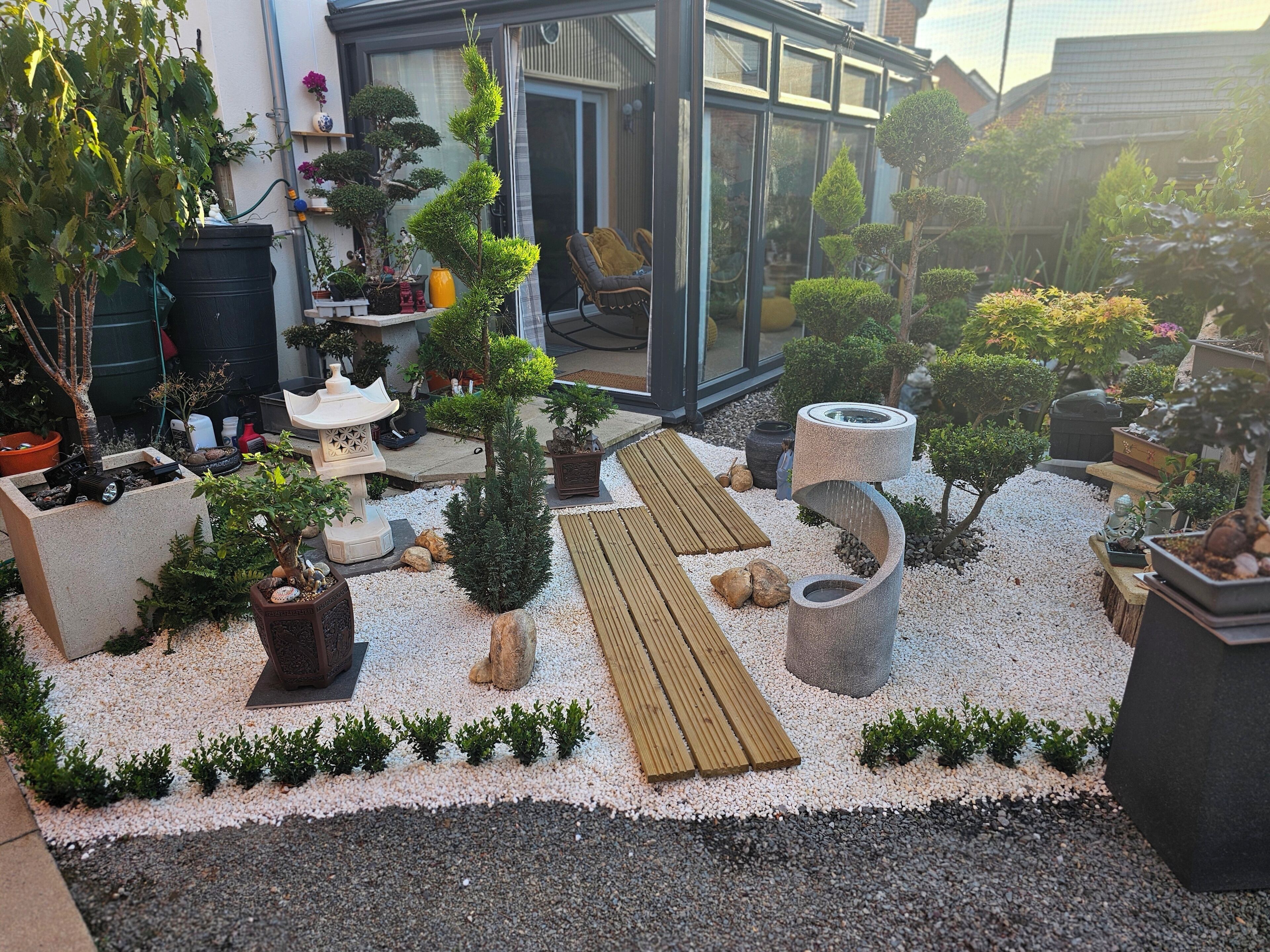 Um jardim bem cuidado com diversos vasos de plantas, um deck de madeira e um elemento aquático de pedra.