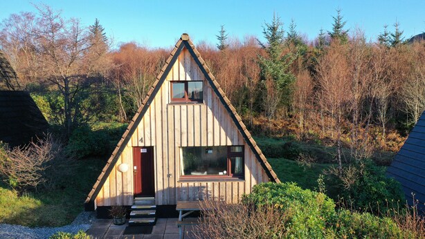 Una cabaña de madera con techo triangular, una puerta principal y un porche pequeño con escalones de acceso.