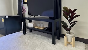 A flat-screen TV on a black TV stand with a remote control, a tissue box, and two glasses on a shelf. A potted plant is on a wooden stand beside the TV. - MONTANA Hotel & Hostel Phuket (Karon)
