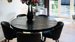 A round dining table with a vase of flowers, surrounded by chairs, in a room with a view through a glass door.