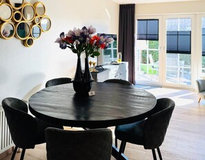 A round dining table with a vase of flowers, surrounded by chairs, in a room with a view through a glass door.
