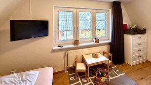 A room with a bed, a TV mounted on the wall, a window with curtains, a radiator, a table with a chair, and a dresser.