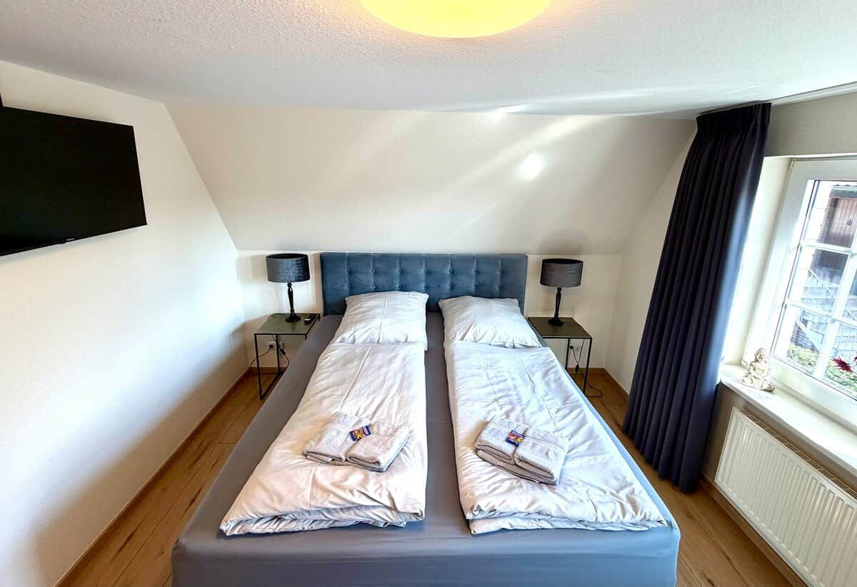 Een tweepersoonsbed met witte lakens, twee nachtkastjes met lampen, een donkerblauwe hoofdbord en een raam met gordijnen.