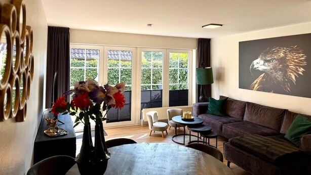 A modern living room with a sofa, coffee table, and chairs, a large window with blinds, and a painting on the wall.