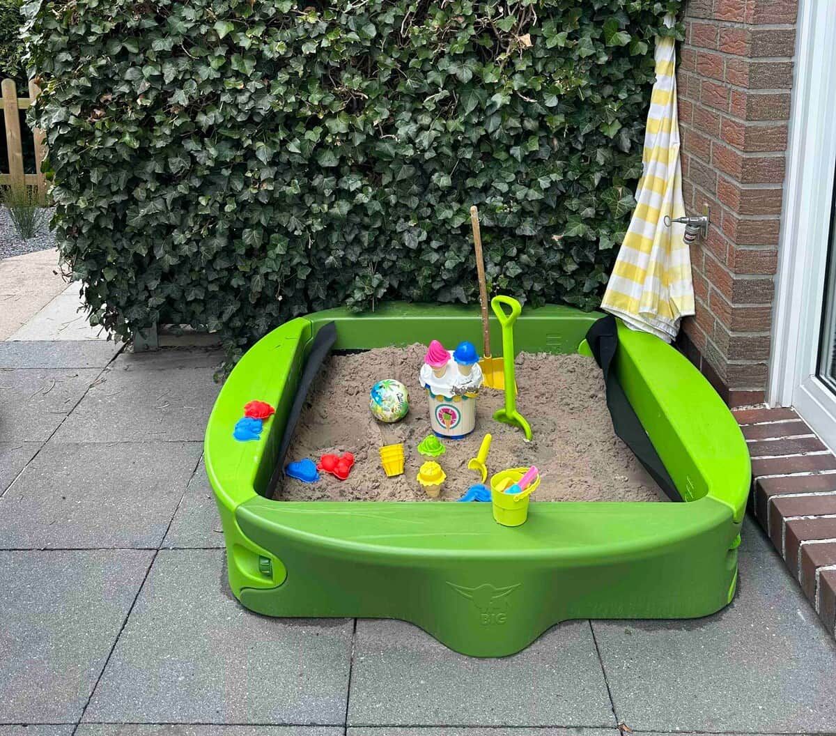 Een groene zandbak met speelgoed en een schep, voor een huis met een bakstenen muur en een houten omheining.
