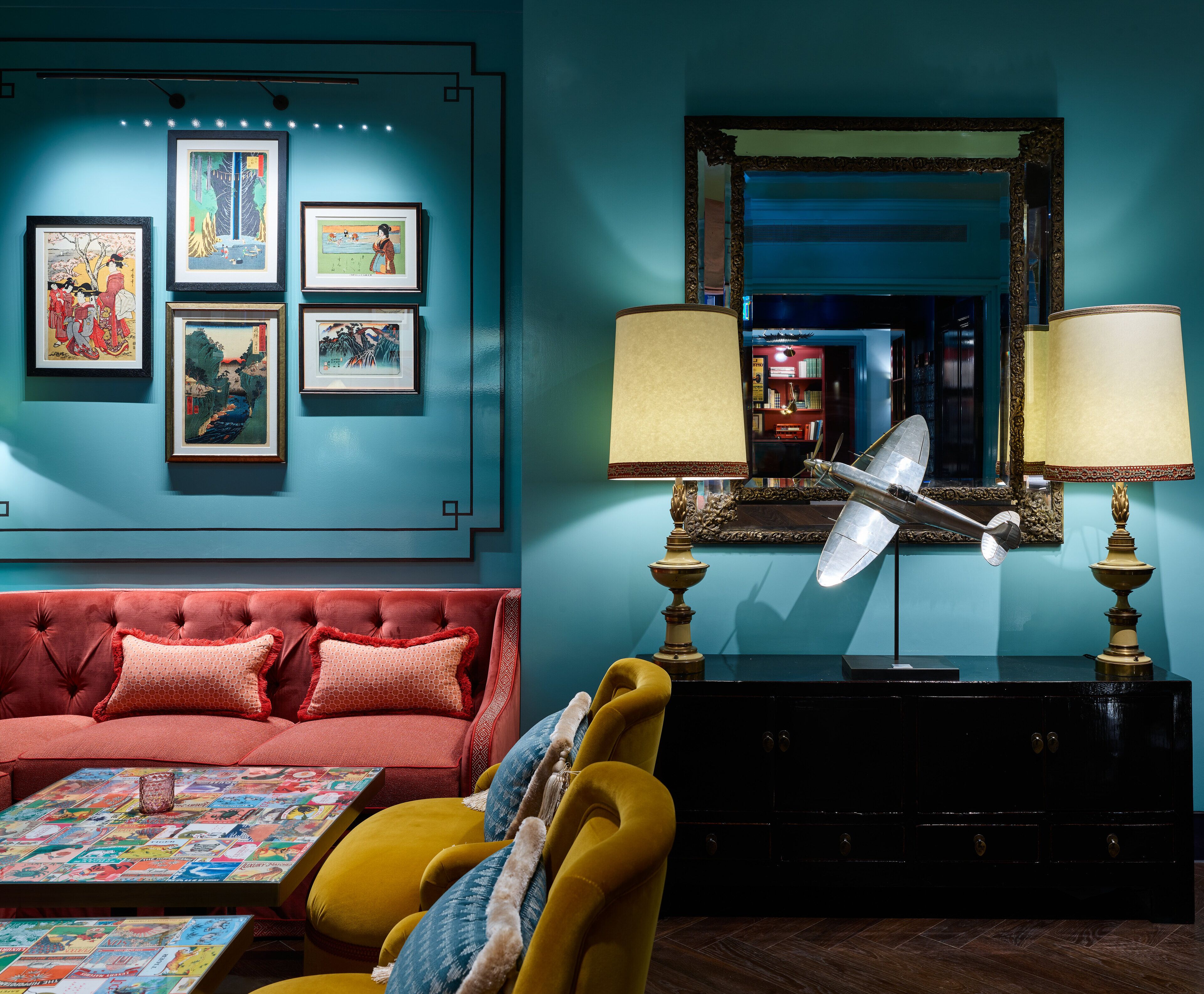 Ein Zimmer mit einer roten Couch, einem gelben Sessel und einem Tisch mit einem farbenfrohen Magazinlayout. An der Wand befinden sich gerahmte Kunstwerke und über einer schwarzen Kommode hängt ein großer Spiegel.