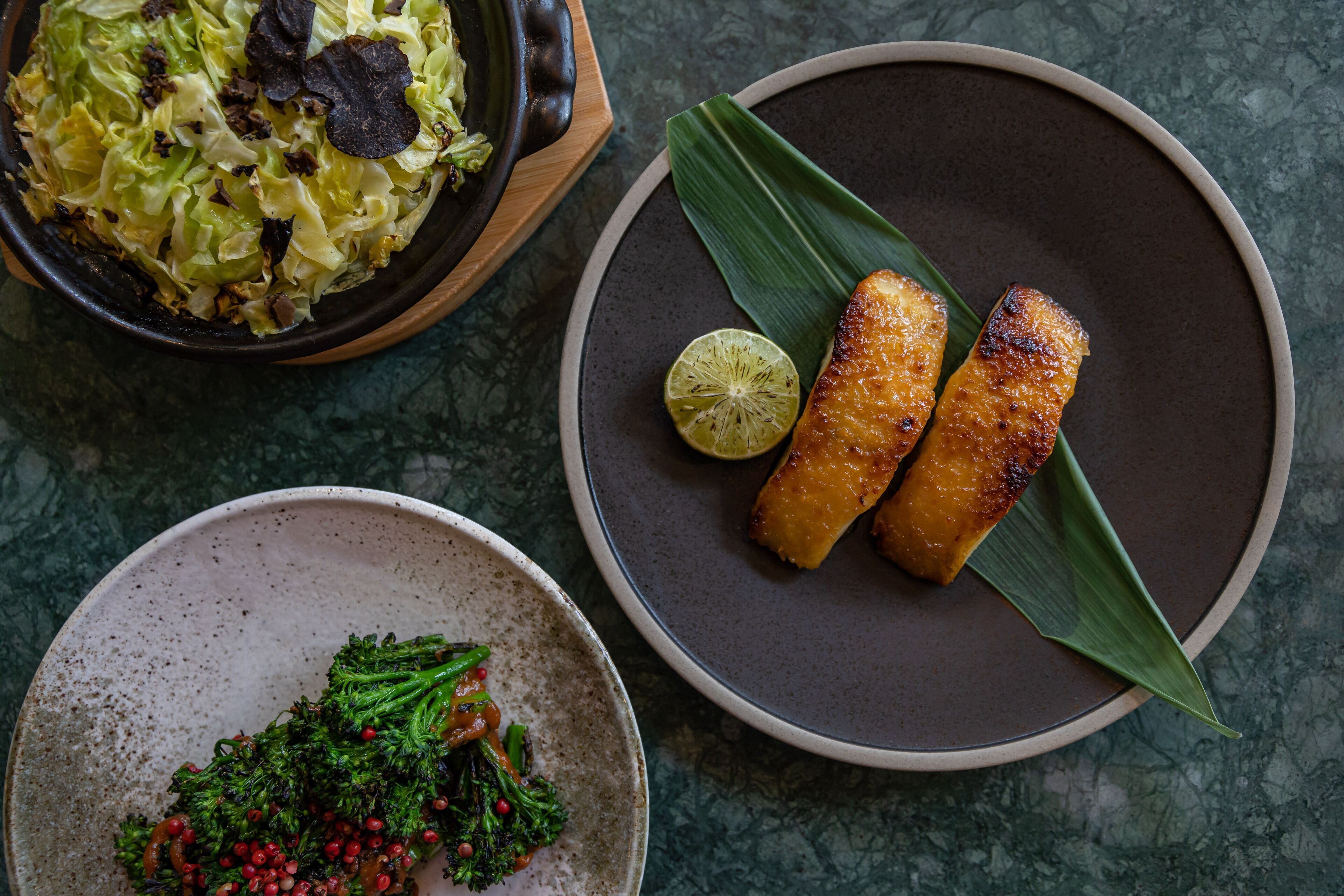 A plate with two pieces of grilled fish, a bowl of cabbage salad, and a plate of broccoli with red peppercorns.