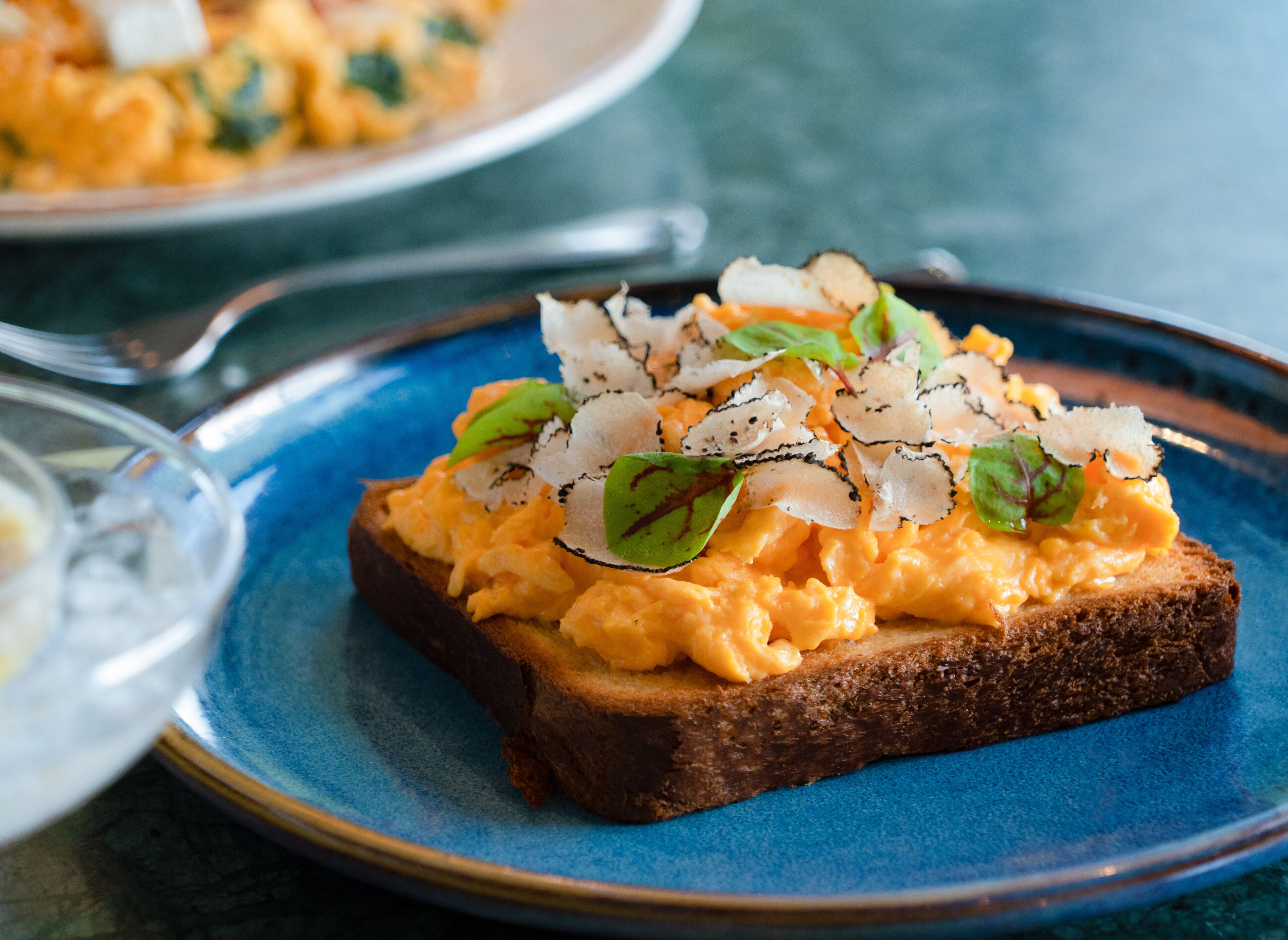A slice of toast topped with scrambled eggs, greens, and shaved cheese on a blue plate.