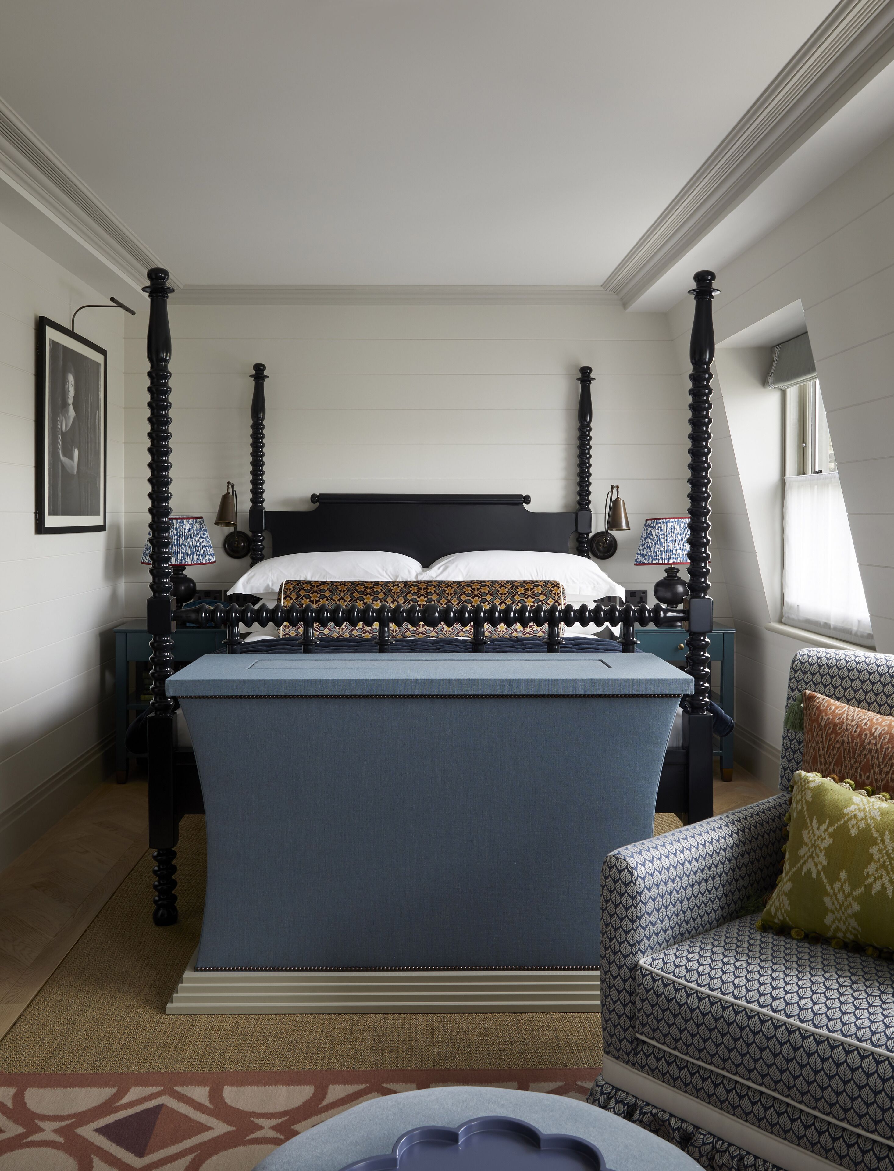 A four-poster bed with a dark headboard, a blue bedspread, and white pillows. There is a patterned armchair with a cushion and a small table with a blue tray.